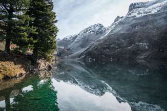 Der Engstlensee ist hier mit besonders klarem, grünlich schimmerndem Wasser zu sehen. Auf der gegenüberliegenden Seeseite befinden sich hohe, felsige Berge mit Schneeresten. Einige Bäume am Ufer spiegeln sich deutlich im ruhigen Wasser.