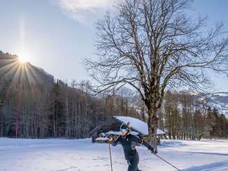 Une femme fait du ski de fond à Grindelwald. Elle est en mouvement, à contre-jour, avec en arrière-plan un arbre dénudé et plusieurs maisons en bois.