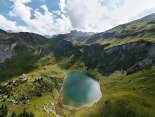 Luftaufnahme des Sulsseeli. Der Bergsee liegt eingebettet in eine weitläufige alpine Landschaft mit grünen Hängen, Felsformationen und Wanderwegen. Die markante Form des Sees hebt sich deutlich von der Umgebung ab.