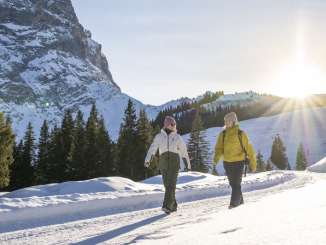 Zwei Personen gehen nebeneinander über eine verschneite Schlittelpiste. Die tief stehende Sonne sorgt für warmes Gegenlicht und lange Schatten im Schnee. Im Hintergrund sind dichte Tannen sowie das Wetterhorn zu sehen. 