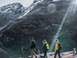 Drei Personen bewegen sich über den Holzsteg mit Ruderbooten. Ein Lichtstrahl fällt von oben ins Bild, während die Berge hinter dem See klar sichtbar sind. Das Bild ist fast identisch zum ersten, nur aus leicht veränderter Perspektive.