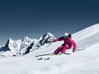 Ein Skifahrer zieht seine Kurven im Skigebiet Mürren-Schilthorn. Das Dreigestirn Eiger, Mönch und Jungfrau ist aus der Ferne zu sehen.