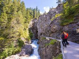 Der Fluss formte die Rosenlaui Gletscherschlucht über Jahrhunderte. Der Weg führt an der Felswand über dem Fluss in die Schlucht hinein. Zwei Wanderer bestaunen den Fluss vor dem Eingang in die Schlucht.