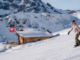 Eine Snowboarderin gleitet die Piste hinunter, während im Hintergrund eine gemütliche Berghütte mit Schweizer Fahne und die eindrucksvolle Alpenkulisse zu sehen sind.