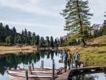 Drei Personen gehen über einen schmalen Holzsteg, an dem mehrere Ruderboote festgemacht sind. Der Engstlensee spiegelt die umliegenden Bäume und den Himmel klar wider. Die Umgebung wirkt herbstlich und ruhig.