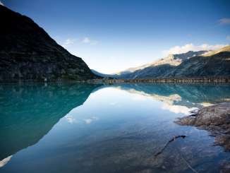 Der Gelmersee liegt eingebettet in der hochalpinen Landschaft der Grimselwelt. Die ruhige Wasseroberfläche spiegelt die umliegenden Bergflanken und den klaren Himmel wider. Im Vordergrund sind die Uferfelsen zu sehen, während die Staumauer den See im Hintergrund begrenzt.
