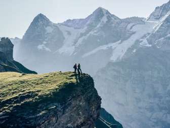 Zwei Wanderer geniessen die Aussicht vom Grauseeli auf einer Klippe. Die Station Birg und das Dreigestirn Eiger, Mönch und Jungfrau strahlen um die Wette.