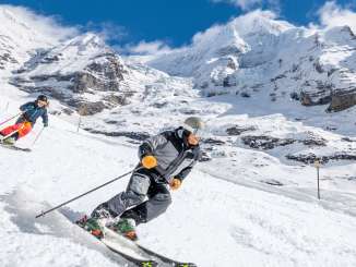 Zwei Skifahrer fahren über eine präparierte Piste. Im Hintergrund ist eine verschneite Bergkulisse zu sehen.