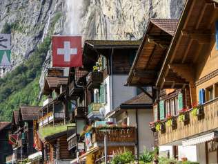 Das Dorf Lauterbrunnen ist umringt von imposanten Felsen. Touristen spazieren durch das Dorf. Im Hintergrund ist der Staubbachfall zu sehen.